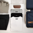 Flat lay of TechTee travel essentials — white, black, and grey classic fit T-shirts neatly folded alongside travel trousers, a packing cube, and carry-on luggage