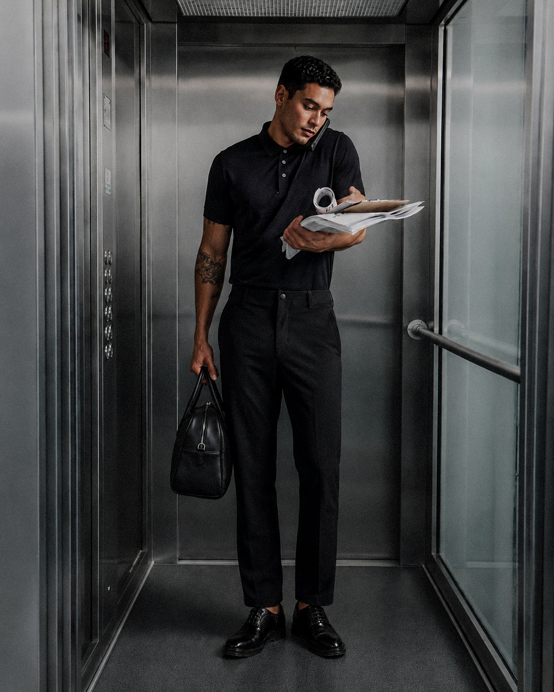 Man in an all black outfit holding a bag and files in an elevator