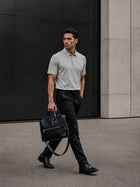 Man in a TECHTEE grey polo shirt and black pants holding a black briefcase, walking against a modern building.